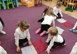 Dzieci siedzą na dywanie i w parach układają z białych i czerwonych chustek flagi